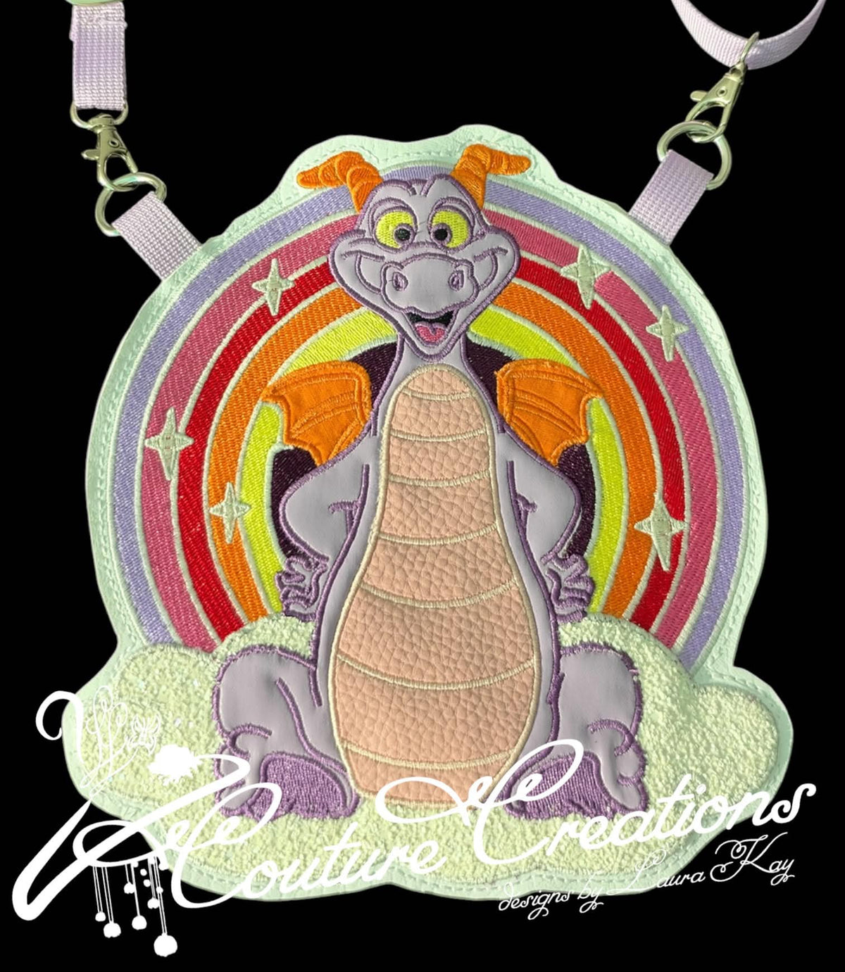 A custom embroidered patch featuring the character Figment, shown on a black background with a lavender strap attached. The patch depicts a smiling purple-gray dragon with orange wings and a peach-colored body, set against a circular rainbow background with white stars.