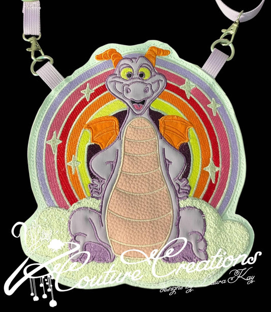 A custom embroidered patch featuring the character Figment, shown on a black background with a lavender strap attached. The patch depicts a smiling purple-gray dragon with orange wings and a peach-colored body, set against a circular rainbow background with white stars.