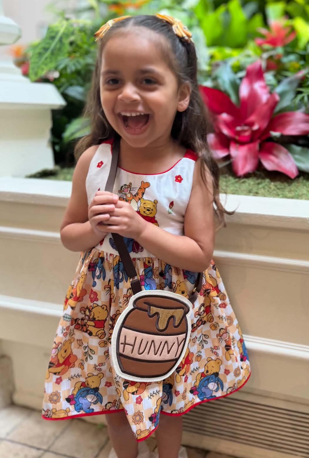 A young girl smiling and laughing while wearing a colorful printed dress and carrying a small shoulder bag shaped like a honey pot with the word 'HUNNY' on it, standing outdoors in front of a planter with green plants and a pink flower.